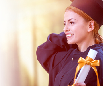 Foto de moça com toga de formatura e barrete segurando um diploma com laço dourado.e olhando para o horizonte.